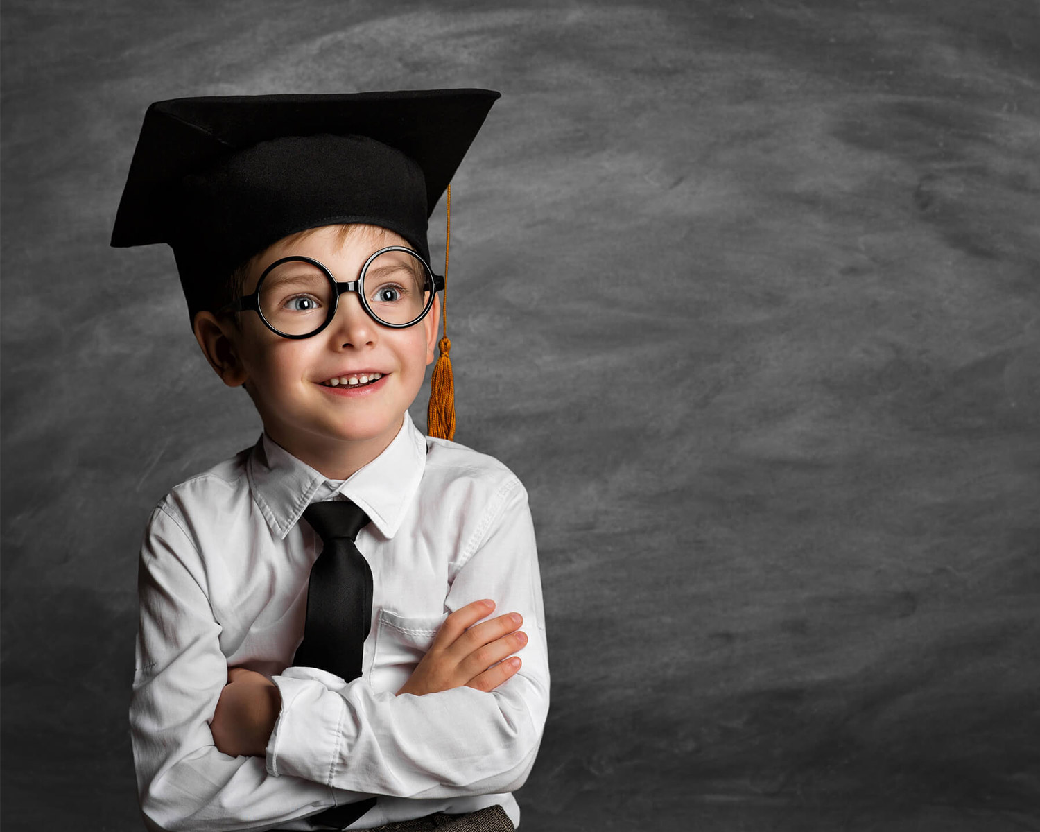 Enfant souriant portant un chapeau de diplômé et des lunettes, bras croisés, devant un tableau noir.