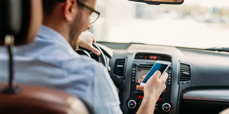 Personne assise au volant d'une voiture, utilisant un téléphone portable dans une main tout en conduisant, illustrant une situation de distraction au volant. 