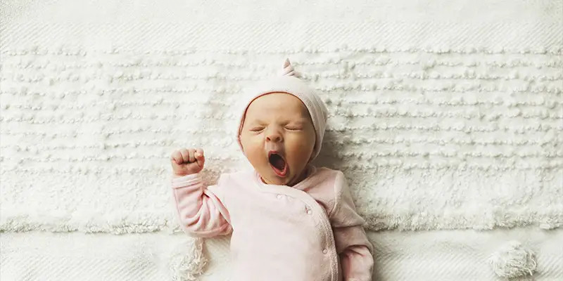 Bébé vêtu d'une tenue rose et d'un bonnet, allongé sur un tissu doux et baillant, les bras levés.