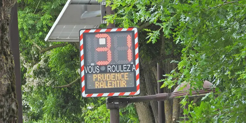 Panneau électronique de signalisation routière affichant "34" et le message "Vous roulez à prudence ralentir", entouré d'une bordure rouge et blanc, installé en extérieur avec des arbres en arrière-plan.