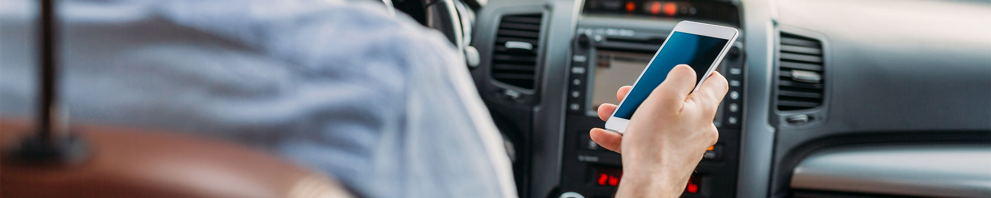 Personne assise au volant d'une voiture, utilisant un téléphone portable dans une main tout en conduisant, illustrant une situation de distraction au volant. 